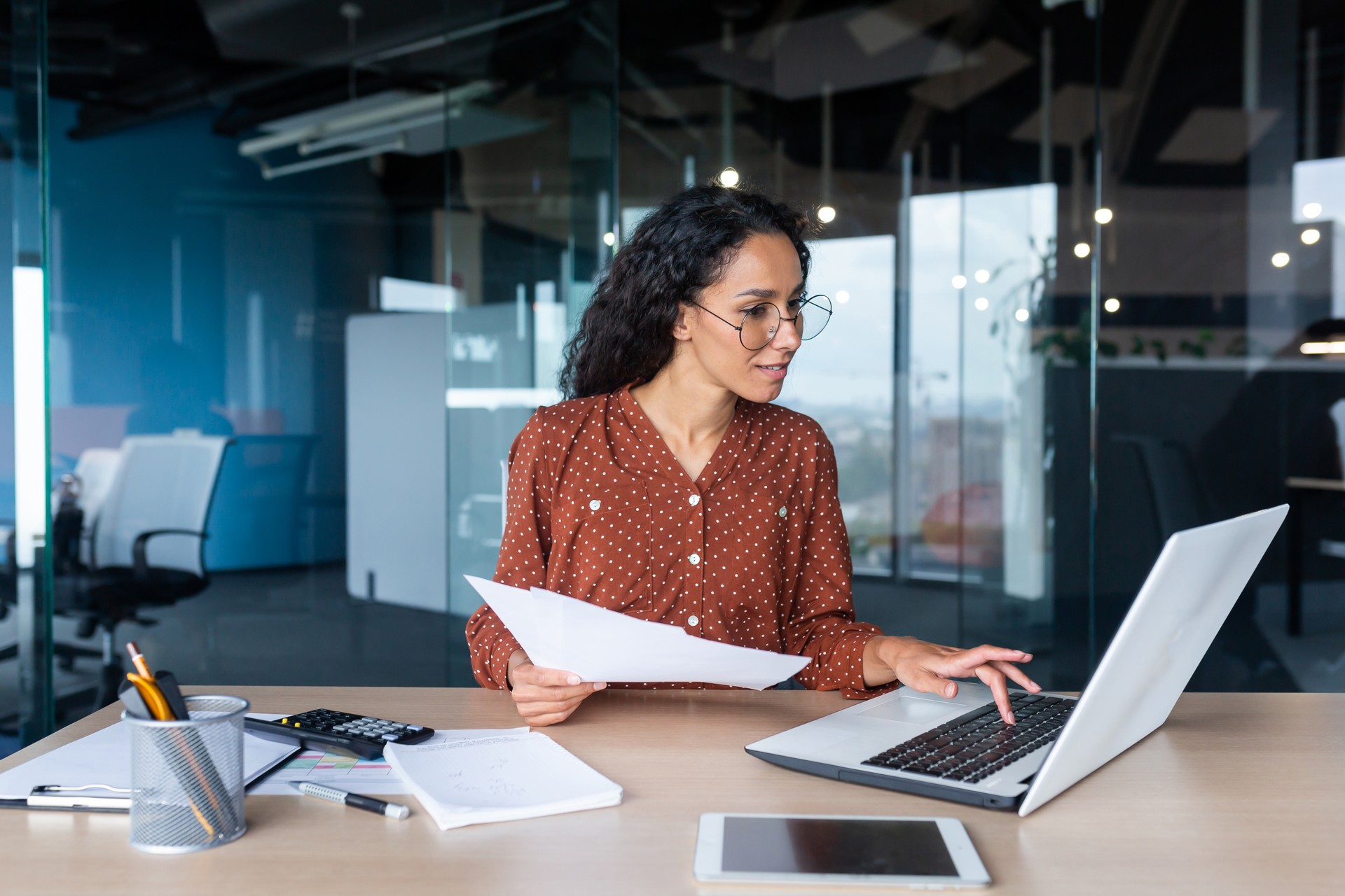 Young happy and successful businesswoman in glasses working with documents inside office, Hispanic woman with laptop looking at bills and contracts, financier with curly hair using laptop