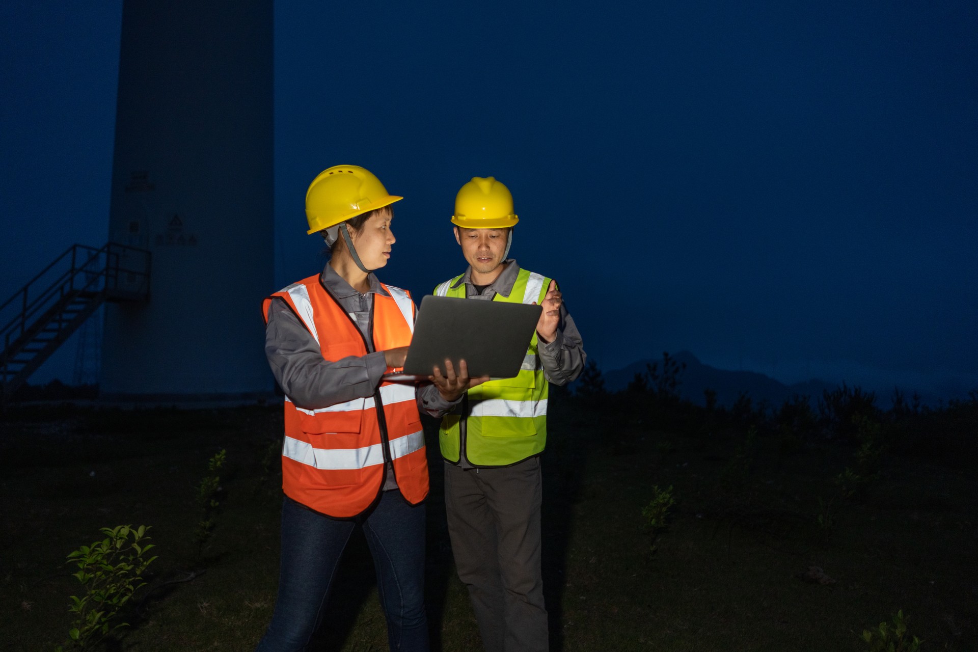 Two Asian engineers on the night shift work with computers under the wind turbine